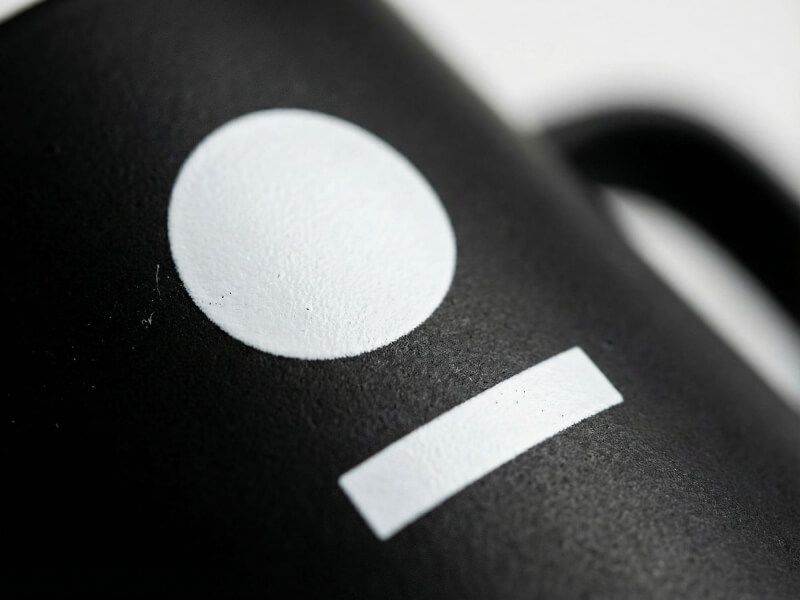 Närbild på en matt svart stengodsmugg med en tryckt vit cirkel och linje, modell Ebba.