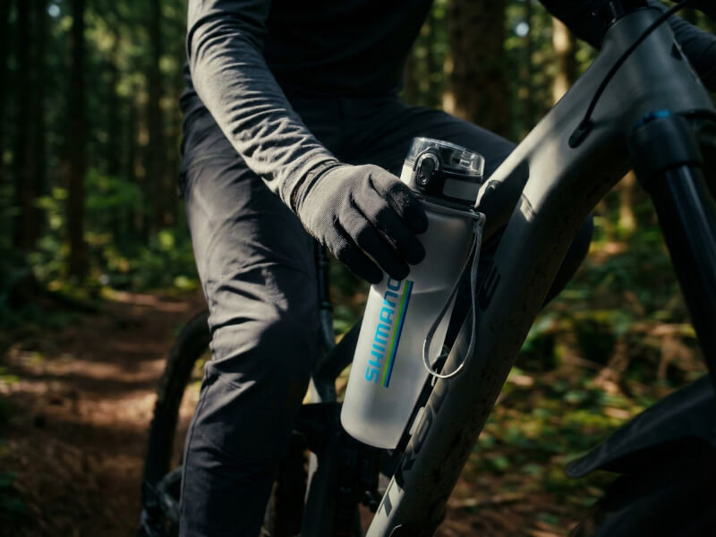 Mountainbikecyklist i helsvart ställ och handskar i en skog som greppar en frostad Shimano sportflaska placerad i ramen på cykeln