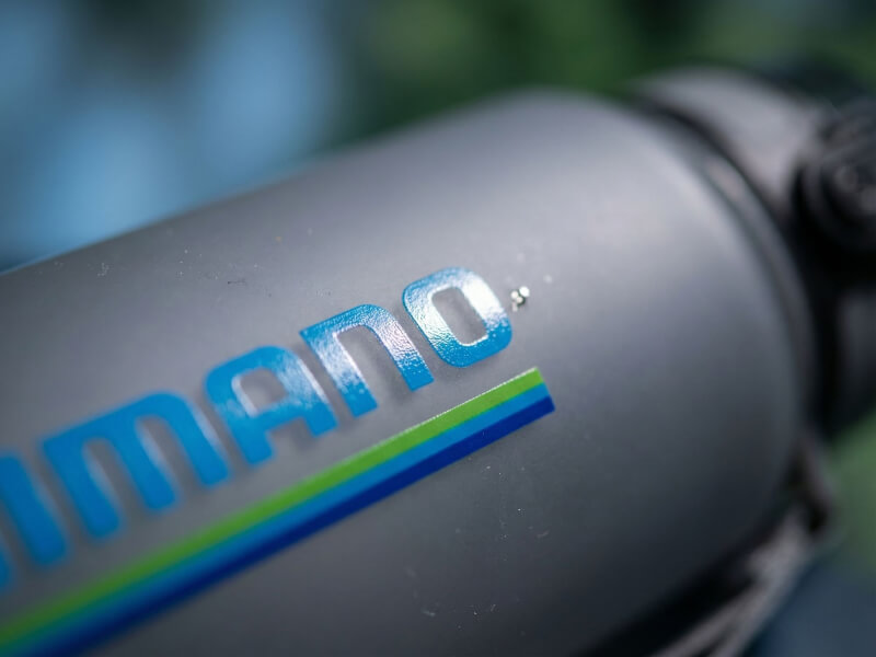 Närbild i vinkel av ljusblå Shimano-logotyp tryckt längs en mörkblå linje på en frostat grå vattenflaska