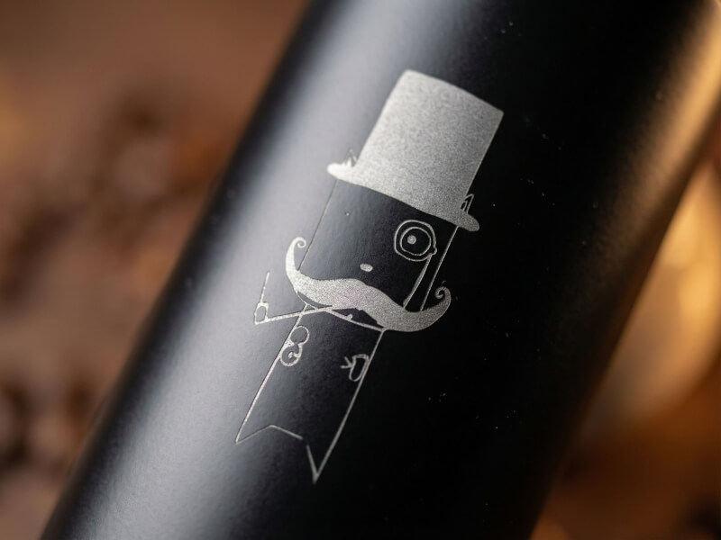 Närbild på en matt svart termosmugg Auto med ett lasergraverat motiv av en figur med cylinderhatt och mustasch, placerad i en mysig miljö.