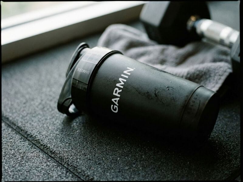 Svart matt Shaker Pro med vit Garmin-logotyp placerad på ett gymgolv bredvid en hantel och träningshandduk.