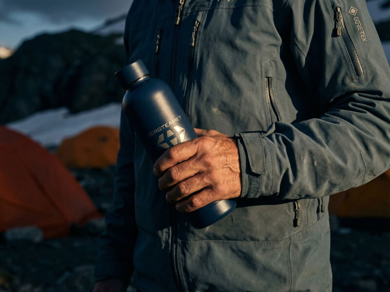 Friluftsklädd person i mörkblå jacka håller tryggt den blå Bergteamet-termosflaskan, med tält uppsatta i bakgrunden