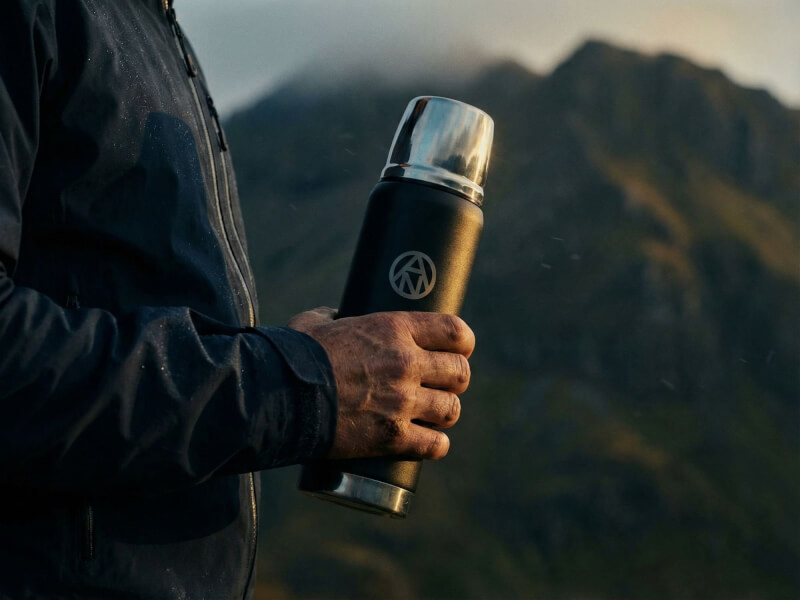 Händer på en person i regnkläder som håller en svart termos med silvergravyr mot en bakgrund av dimmiga, branta berg och vild natur