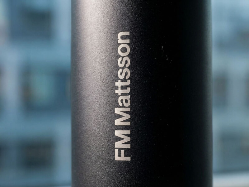 Oskarp närbild på vertikalt ingraverad, silverfärgad FM Mattsson-logotyp i stilrent typsnitt på en mörk termos