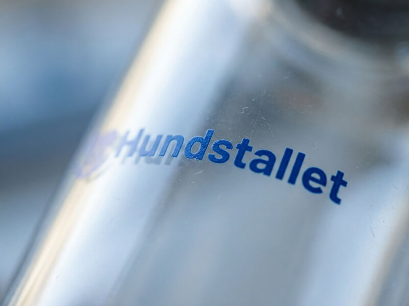 Urgjort kort skärpedjup av en liten blå logotyp för Hundstallet tryckt på mitten av en slät glasvattenflaska