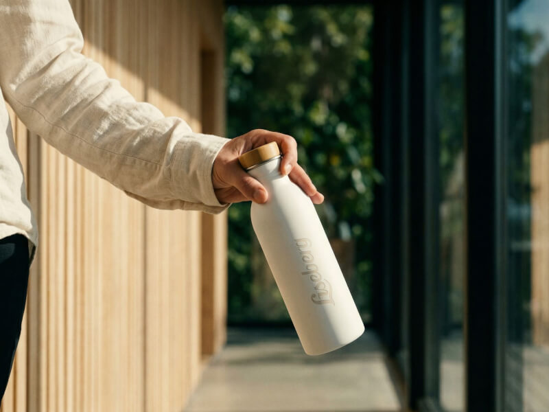 Person som sträcker sig med en vit Liseberg-vattenflaska framför en modern träfasad i eftermiddagssol