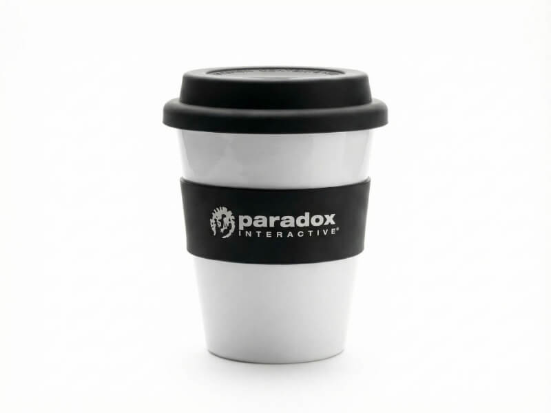 Vit plastmugg Astoria med svart silikongrepp och svart lock, profilerad med Paradox Interactive-logotyp genom tryck, mot vit bakgrund.