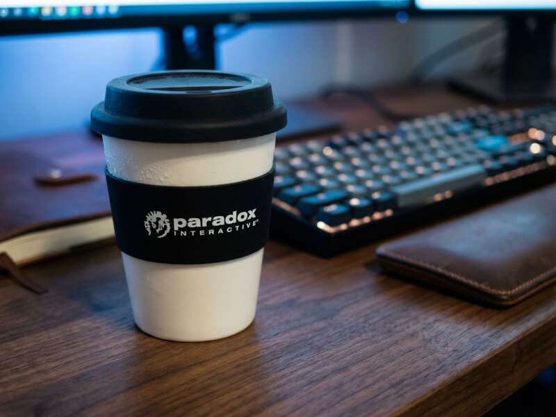 Vit plastmugg Astoria med svart silikongrepp och lock, placerad på ett skrivbord i en kontorsmiljö med en Paradox Interactive-logotyp tryckt på greppet.