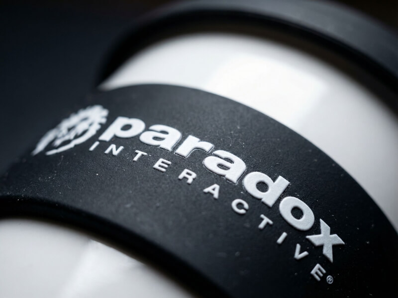 Närbild på Plastmugg Astoria med vit blank yta och svart matt silikonband märkt med Paradox Interactive-logotyp i vitt tryck.