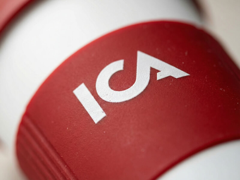 Närbild på en vit plastmugg Americano Grip med ett rött greppvänligt silikonband och ICA-logotyp tryckt i vitt.