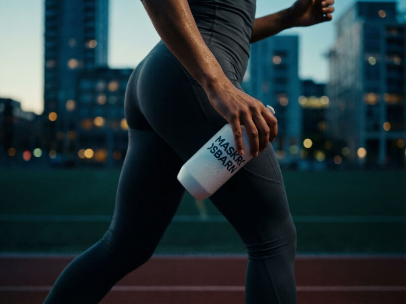 Person klädd i mörka träningskläder som joggar utomhus kvällstid i en stadsmiljö, hållandes en mattvit Maskrosbarn-sportflaska