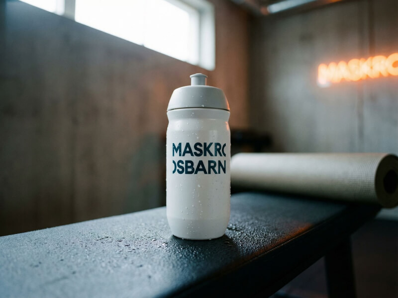 Vit Maskrosbarn-sportflaska stående på en svart träningsbänk i ett fitness-studio upplyst av neonljus