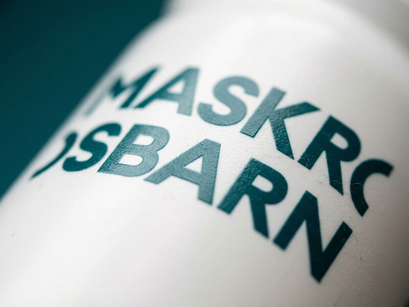 Utökad närbild med dramatisk vinkel av textlogotypen Maskrosbarn tryckt i mörkgrönt på en mattvit sportflaska
