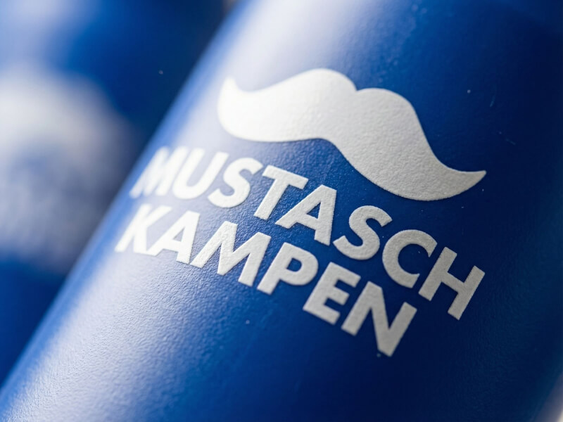 Snäv närbild vinklad uppåt på vit mustasch-ikon och texten Mustaschkampen tryckt på en blå sportflaska