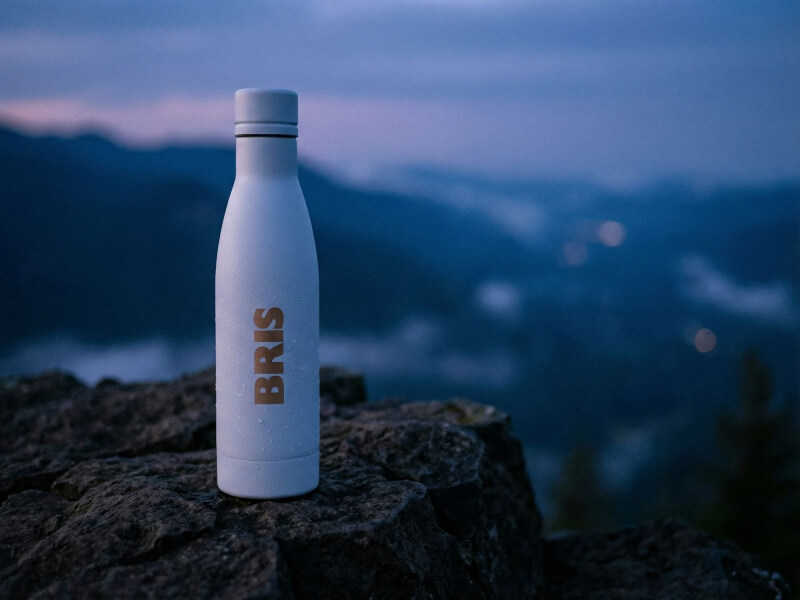 Vit matt termosflaska Vasa Återvunnen med vattendroppar och bronsfärgat logotryck, placerad på en klippa med en mörk bergsmiljö i skymning i bakgrunden.