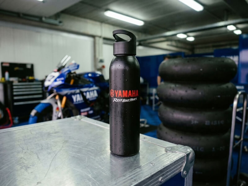 Svart stålvattenflaska med Yamaha-tryck stående på ett grått bord inuti ett välbelyst garage med motorcyklar och däck i bakgrunden